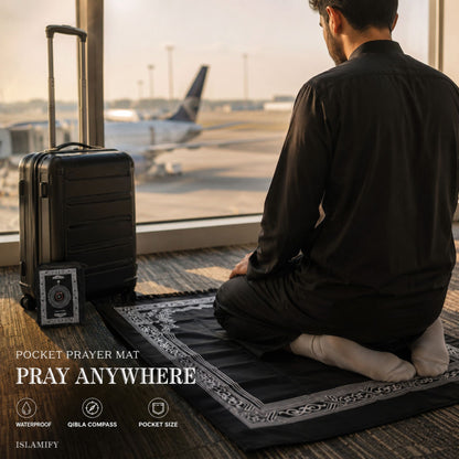 A man in a black shirt and pants sits on a black prayer mat with a decorative border, facing a large window that shows an airplane on the tarmac. A black hard-shell suitcase stands beside him, and a small prayer compass is attached to the suitcase.