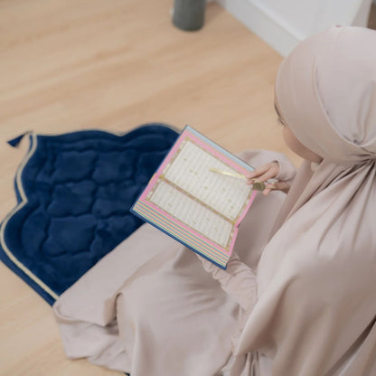 Muslim woman reading Quran on a navy foam padded prayer mat by Islamify in a serene setting